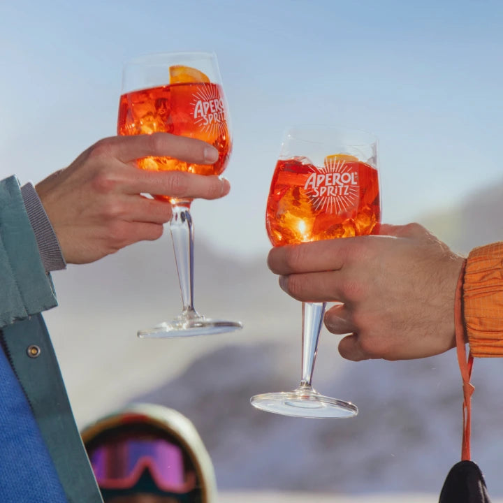 Two hands toasting with Aperol Spritz glasses against a snowy mountain backdrop, evoking a festive après-ski vibe.